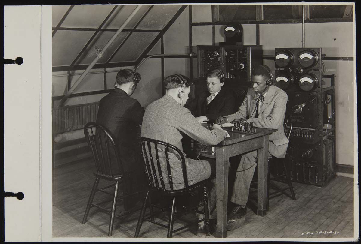Four young men wearing headsets sit at a table with machinery, with additional machinery behind them