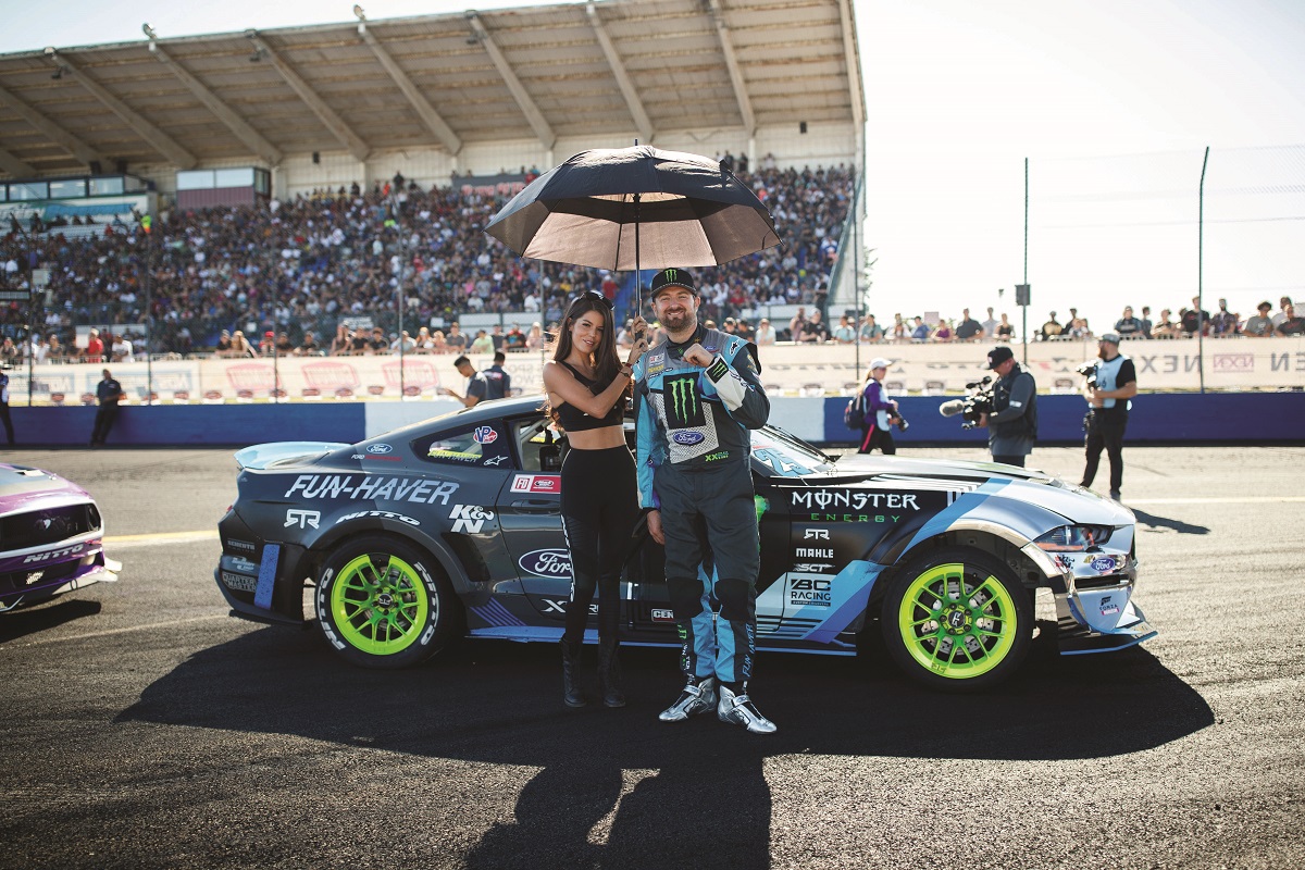 Woman holding umbrella stands with man in racing jumpsuit in front of racecar on track with spectators in grandstands behind them
