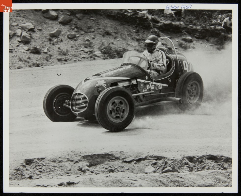Person wearing face mask, helmet, and jumpsuit drives race car on dirt road with rocky slopes on either side