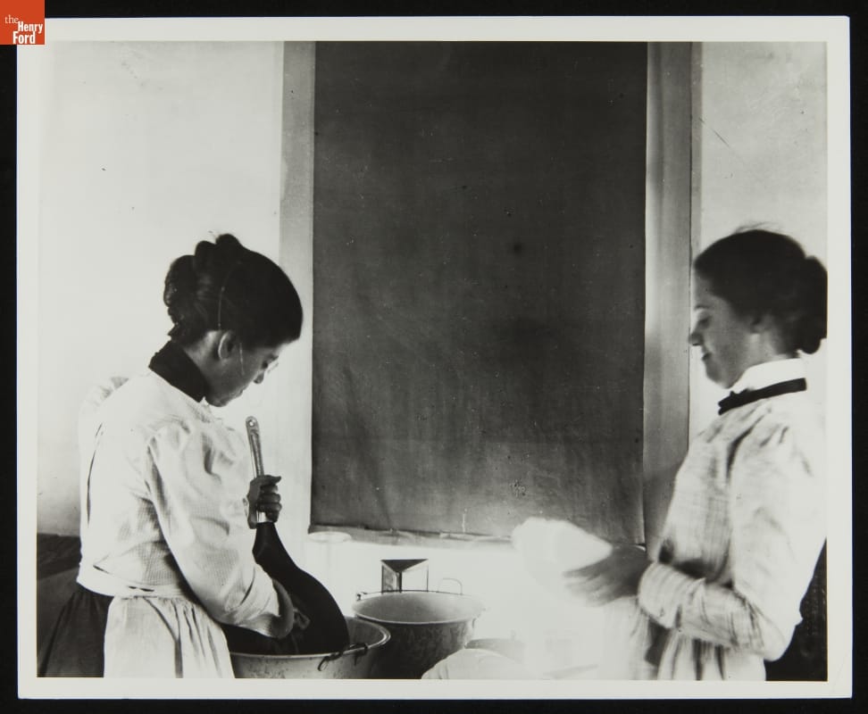 Two women wash dishes among tubs of water in front of a window with a closed blind