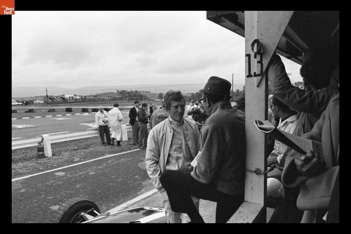 Two men talking; other people in foreground to right and in background are more people and a raceway