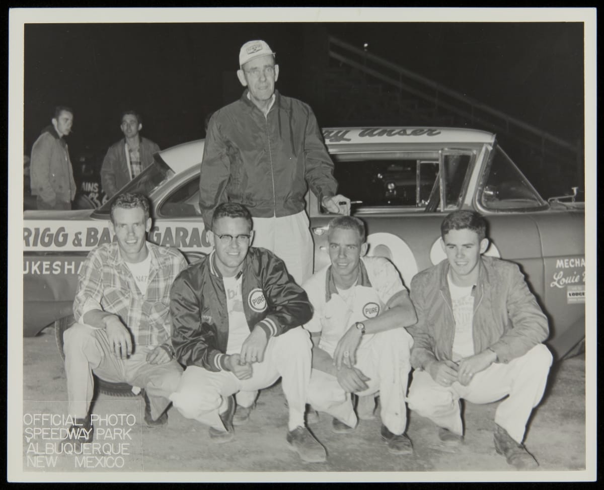 Black-and-white photo of man standing in front of racecar, with four younger men kneeling in front of him