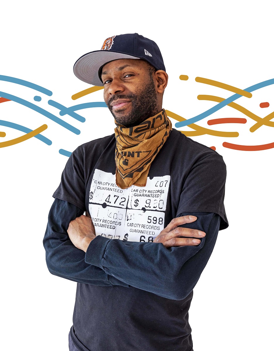 African American man wearing long-sleeve t-shirt, bandana around neck, and baseball cap stands with arms crossed with a colorful pattern of wavy lines in the background