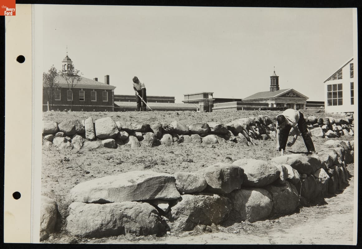 Two young men or boys work in tiered garden plots divided by boulders with buildings in the background