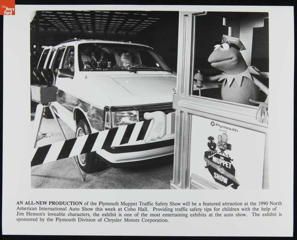 Muppet Traffic Safety Show Sponsored by Plymouth, North American International Auto Show, 1990 Van containing Muppet characters stopped behind barrier; Kermit the Frog in a booth in foreground; contains text