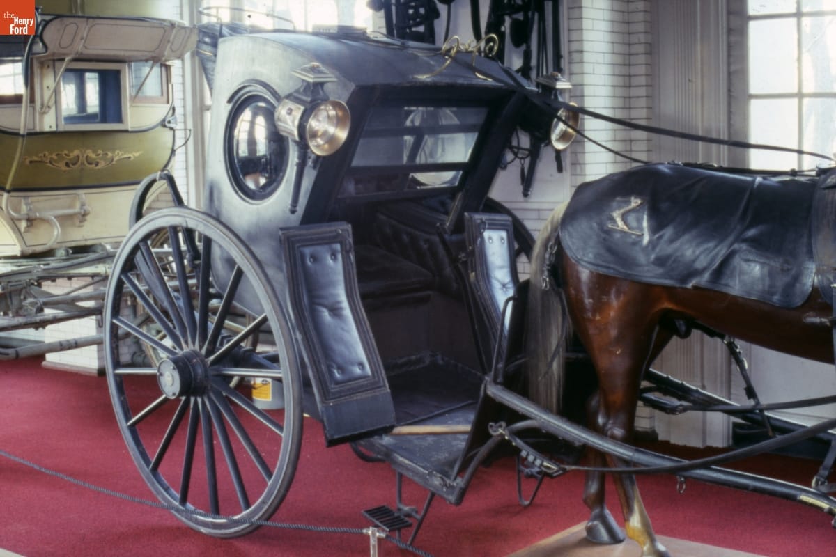Two-wheeled black carriage on display with other vehicles and horse mannequin