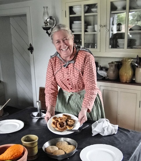 A woman in a gingham dress and apron smiles and holds a plate of food in a kitchen