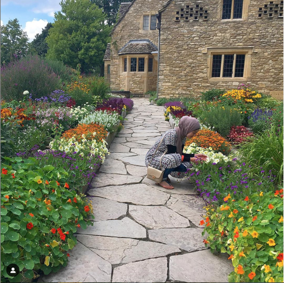 A woman kneels in the garden at Cotswold Cottage in Greenfield Village. Woman in a dress and hijab kneels on a gravel path through a flowerbed with a beige stone cottage in the background