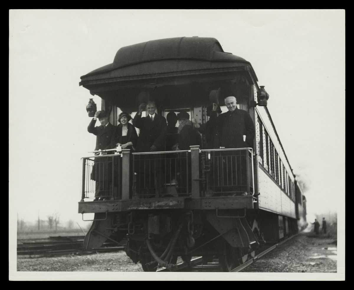 Black-and-white photo of six people on the back platform of a rail car, most of them smiling and waving