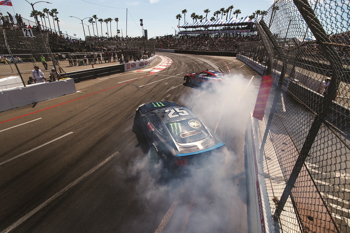 Two race cars drift and spew smoke on a racetrack surrounded by fencing, palm trees, and spectators in stands