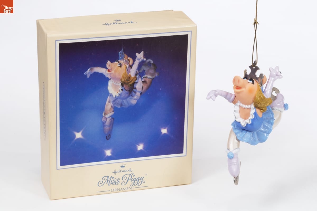 Ornament of Miss Piggy in blue-and-white ice skating costume with long white gloves, arms and legs in dramatic pose, sitting next to box with image of the same ornament on the front