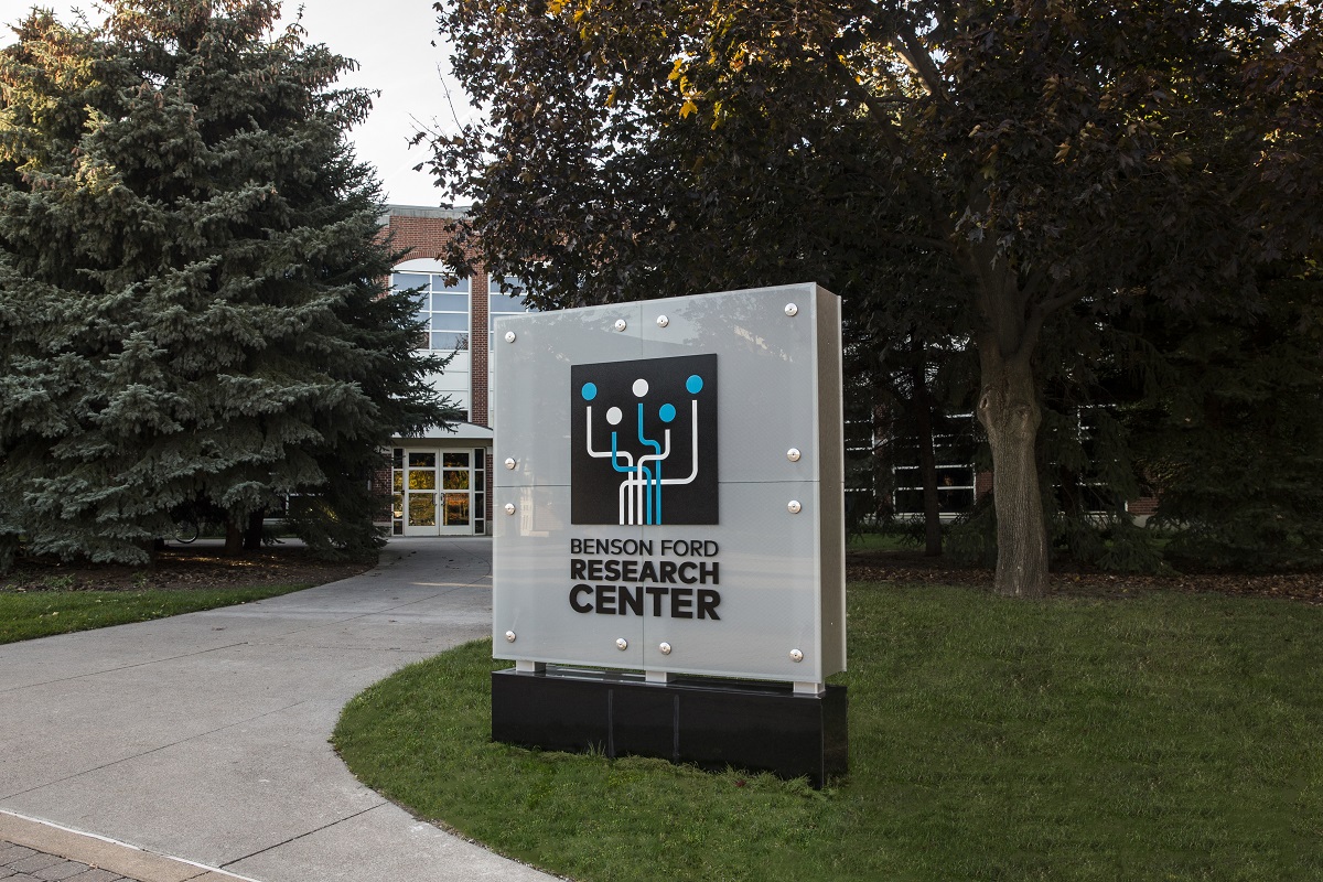 The entrance to the Benson Ford Research Center at The Henry Ford Photo of gray sign with text and logo on lawn next to curving sidewalk leading to a brick building