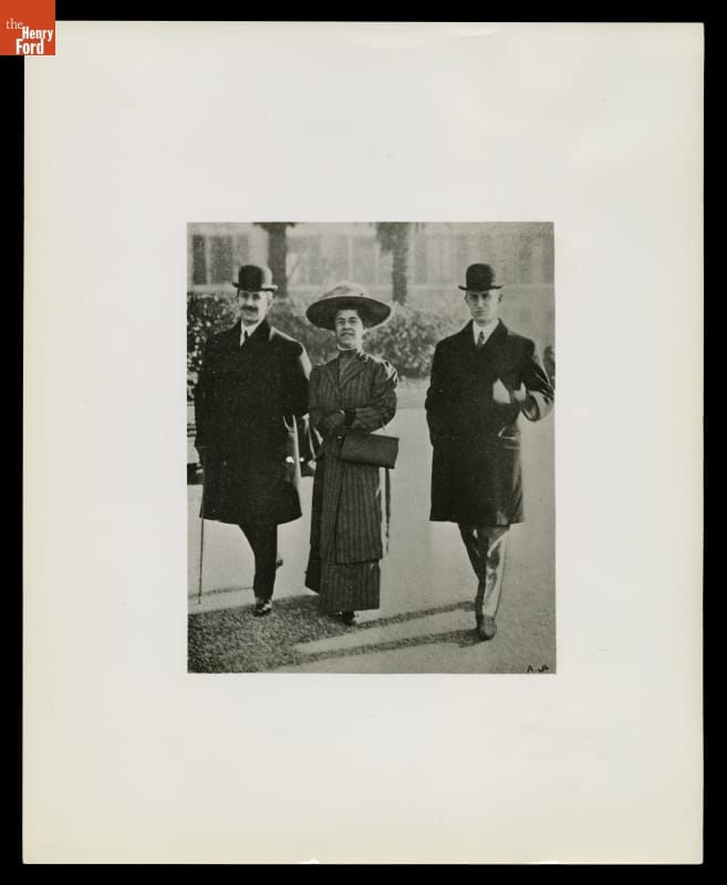 Matted photograph of woman in dress and hat walking between two men in coats and bowlers