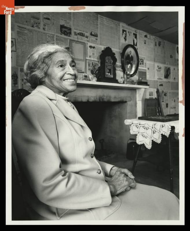 Rosa Parks Visiting Mattox House in Greenfield Village, 1992 Woman with hair pulled back wearing suit jacket sits in chair with mantel and wall covered in newspaper behind her