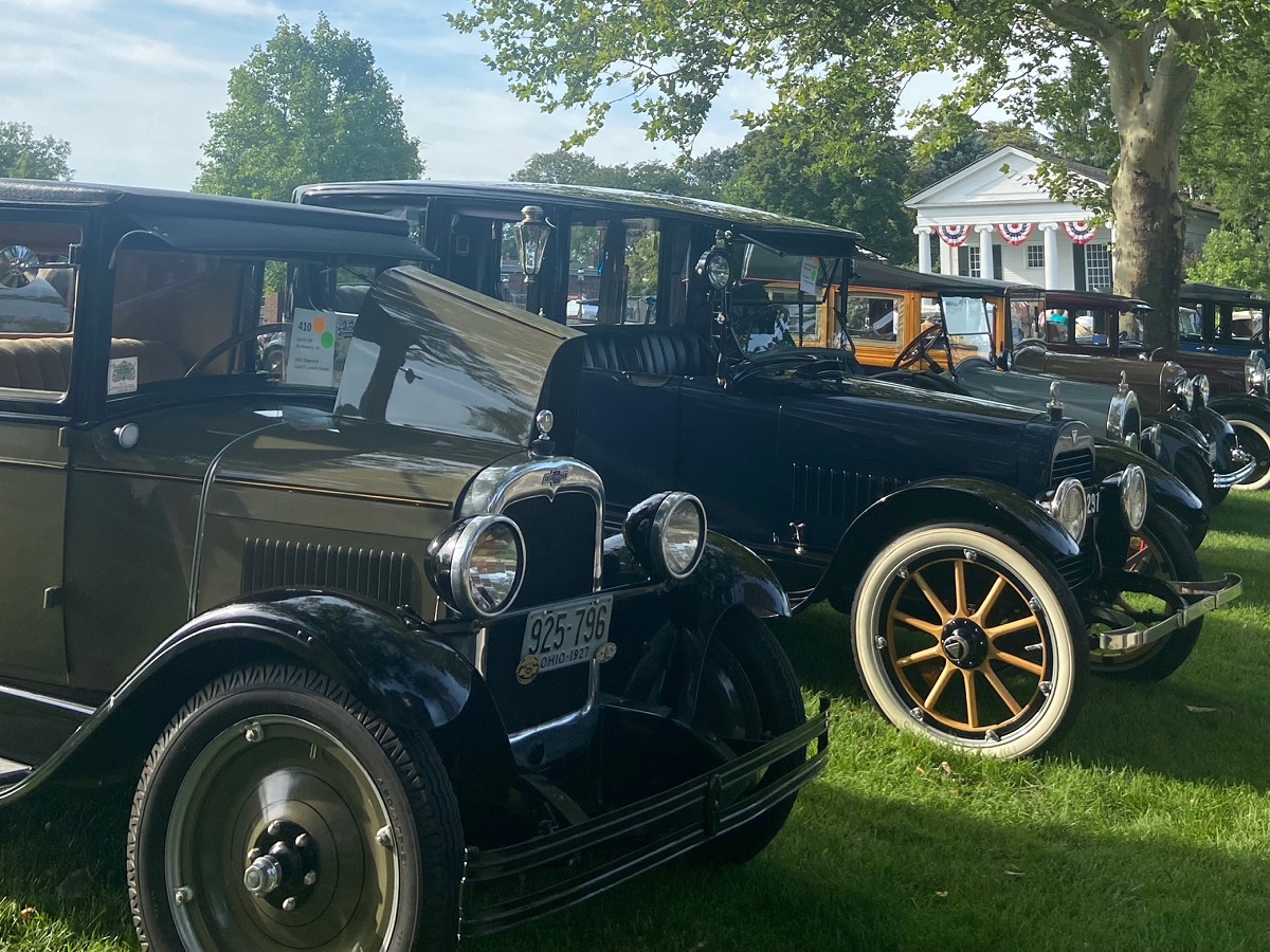 A row of old-fashioned cars are parked on a green lawn in front of a white building with columns decorated with red, white, and blue bunting