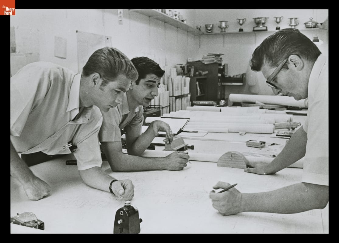 Three men lean over a large table filled with drawings and other items