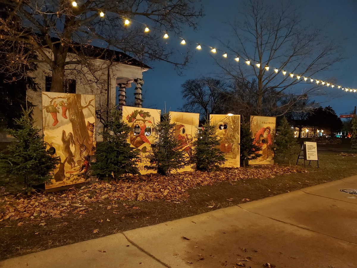 Life-sized postcards at Holiday Nights in Greenfield Village A row of large wooden backdrops with holes cut for people's faces stands in front of a building along a sidewalk