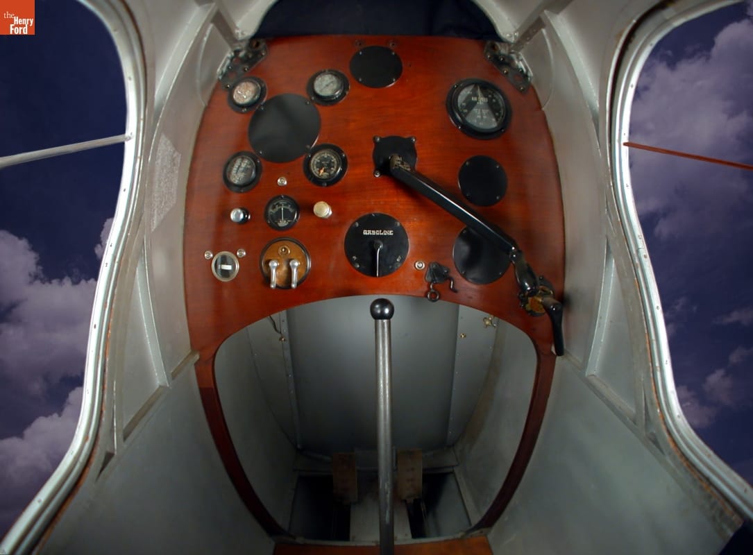 Plane cockpit with large wooden panel of instruments and gauges above legroom with pedals; no windows in front but large windows to the left and right