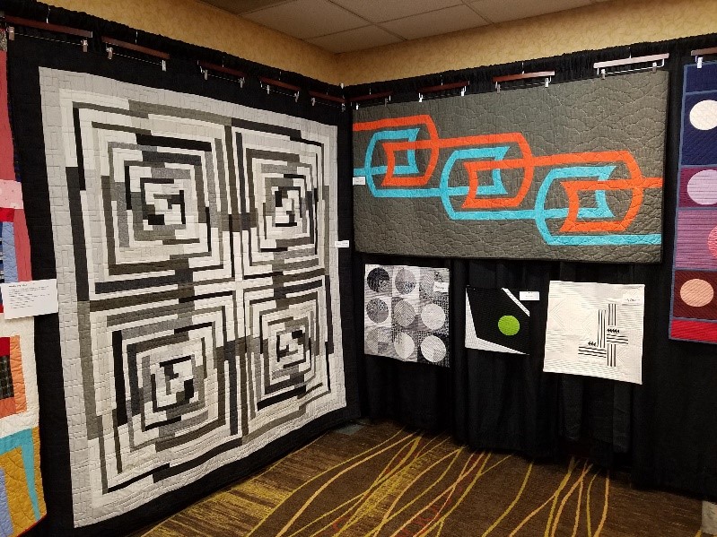 Quilt display at Lincoln, Nebraska Modern Quilt Guild at American Quilt Study Group’s October 2019 Seminar Quilts, mostly shades of black, white, and gray, hanging on a black backboard
