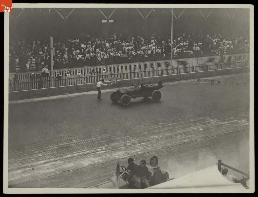 Black-and-white photo of an old-fashioned race car on a dirt track in front of stands filled with fans