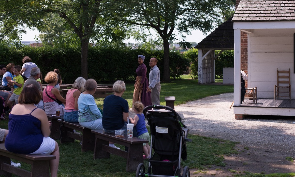 Anthony Lucas in "How I Got Over" Man and woman stand in front of white wooden house as a group of people seated on benches watch