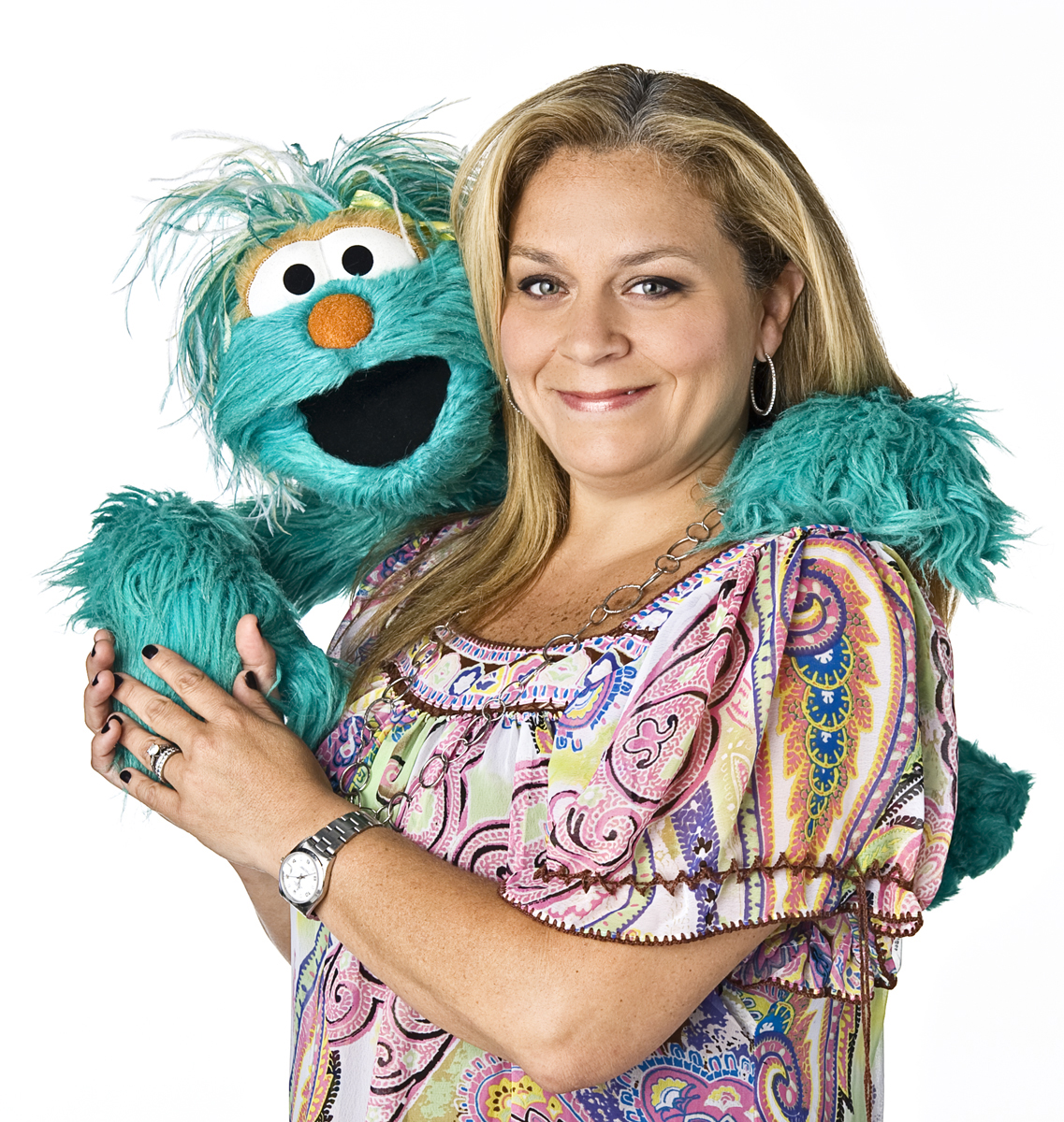 Smiling woman with long blonde hair stands with blue furry puppet over one shoulder