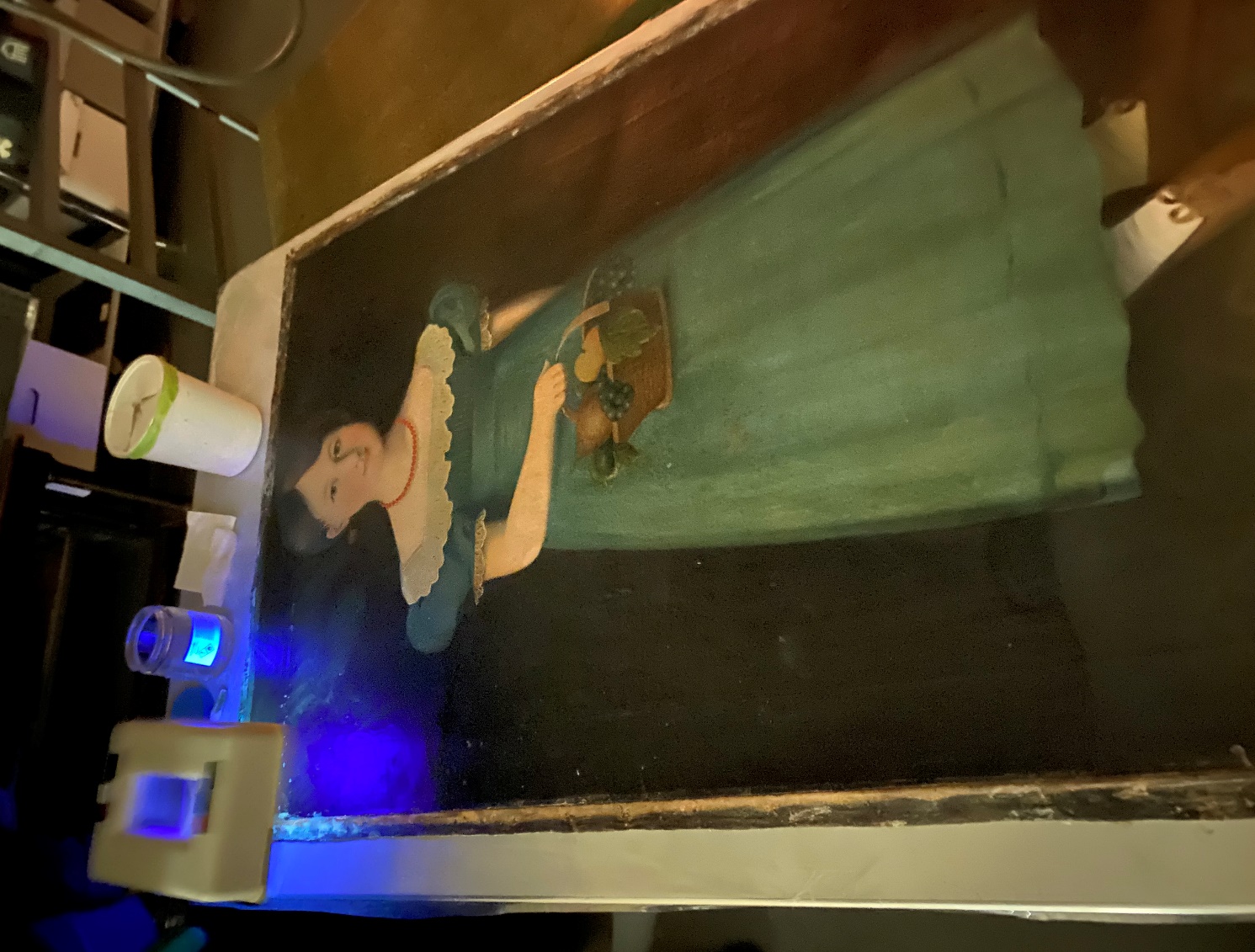 Painting of girl in blue dress, laid flat on table with blue light and tools at one end