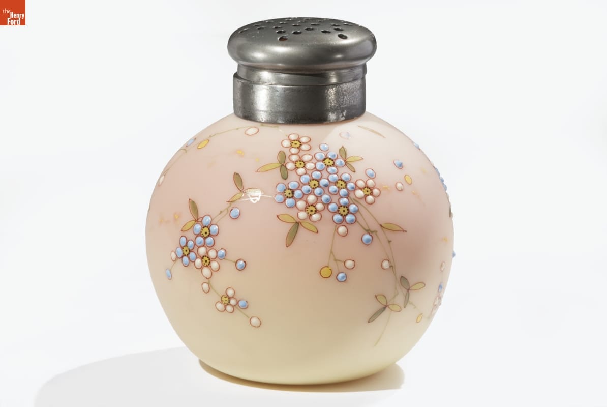 Round glass container with "salt shaker" type top and raised pattern of flowers, in shades of cream and peach