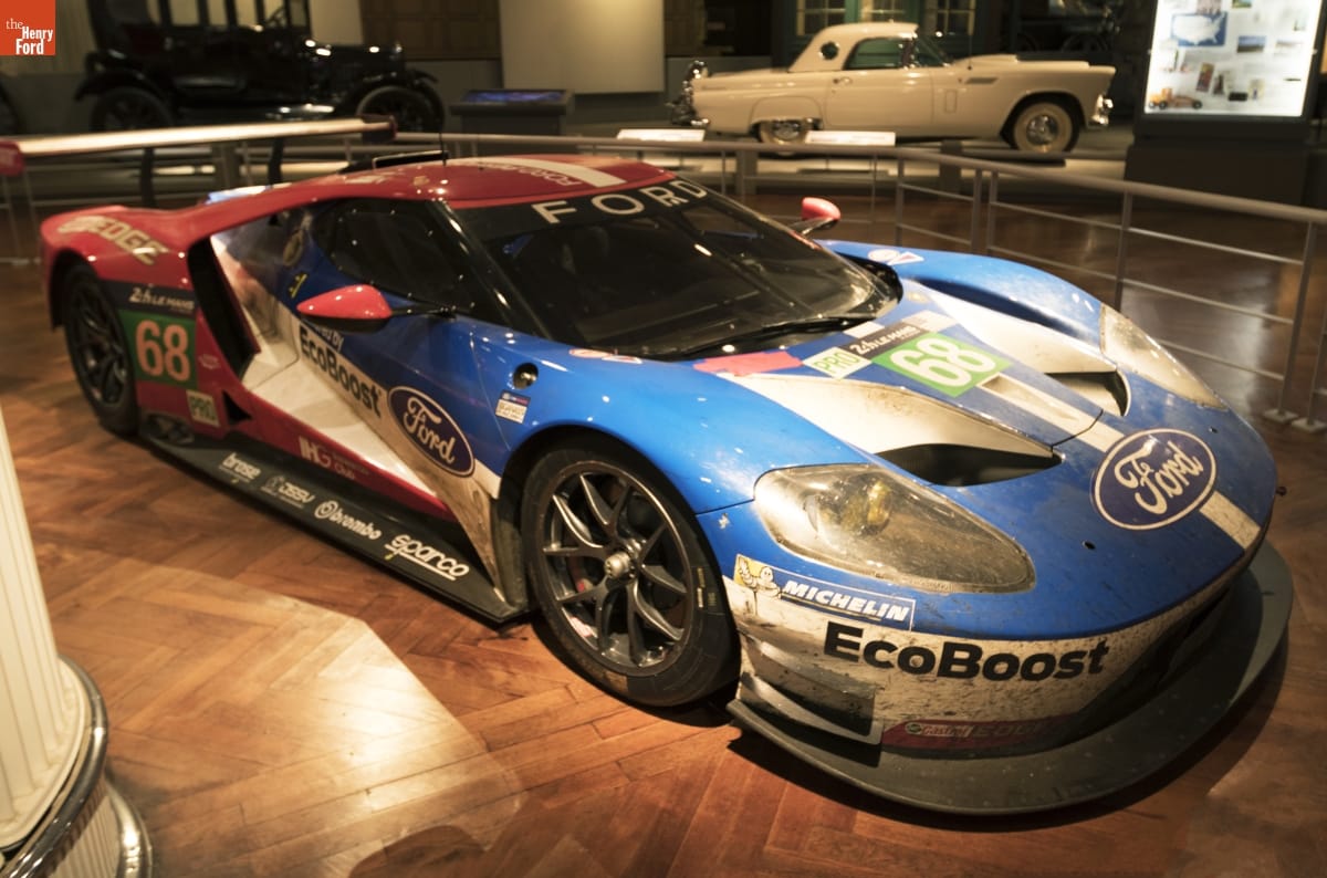 Low blue, red, and white race car with text and logos, sitting in a large indoor space with other cars nearby
