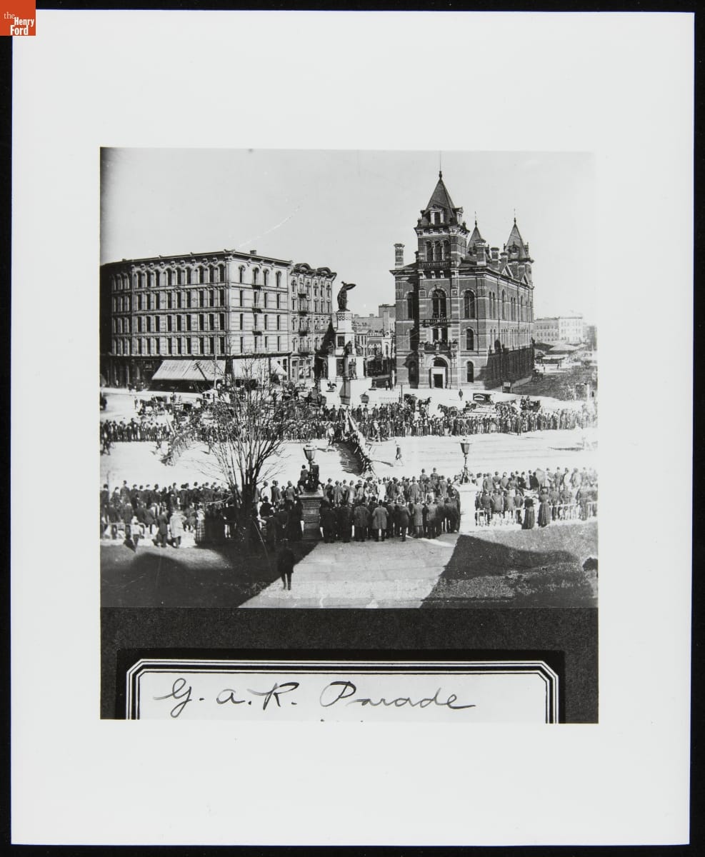Open city square containing large monument and many people gathered around