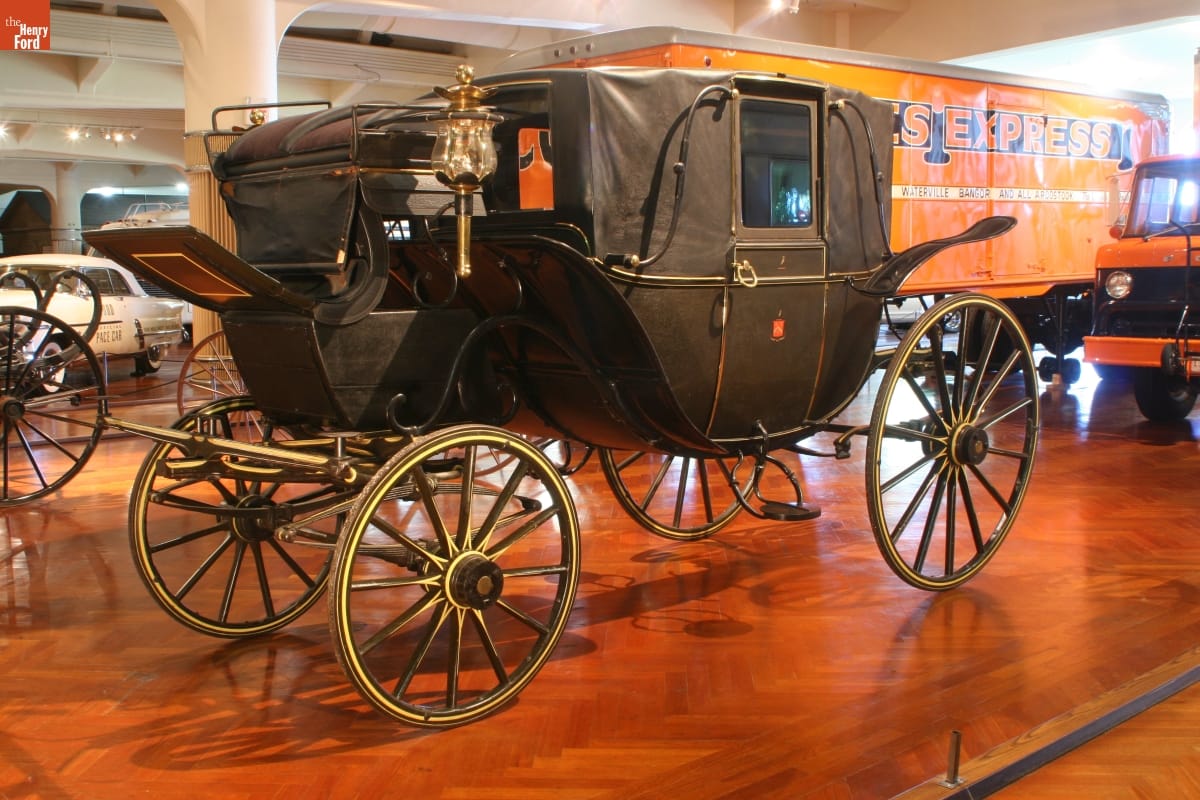 Black carriage with elevated coachman's seat in front; in room with other vehicles of various types