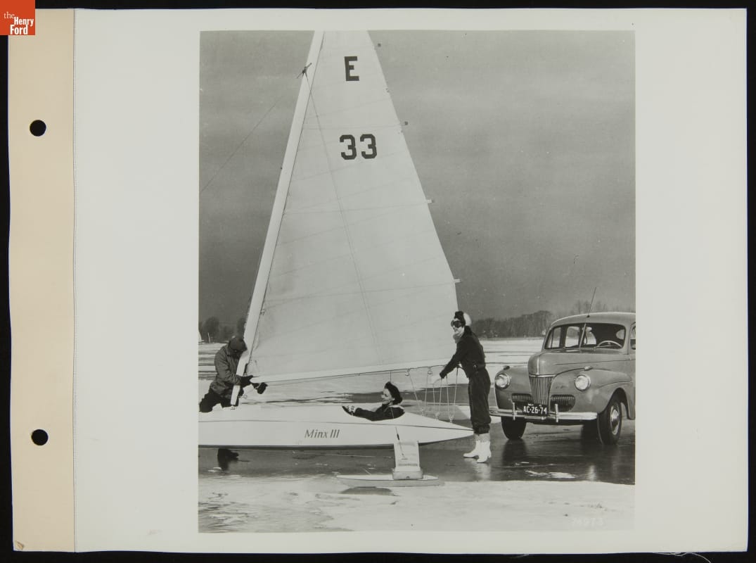 Woman sits in a long, low conveyance with sail and “Minx III” printed on side, on a bed of ice and snow, while two more women adjust the sail and a car is parked nearby