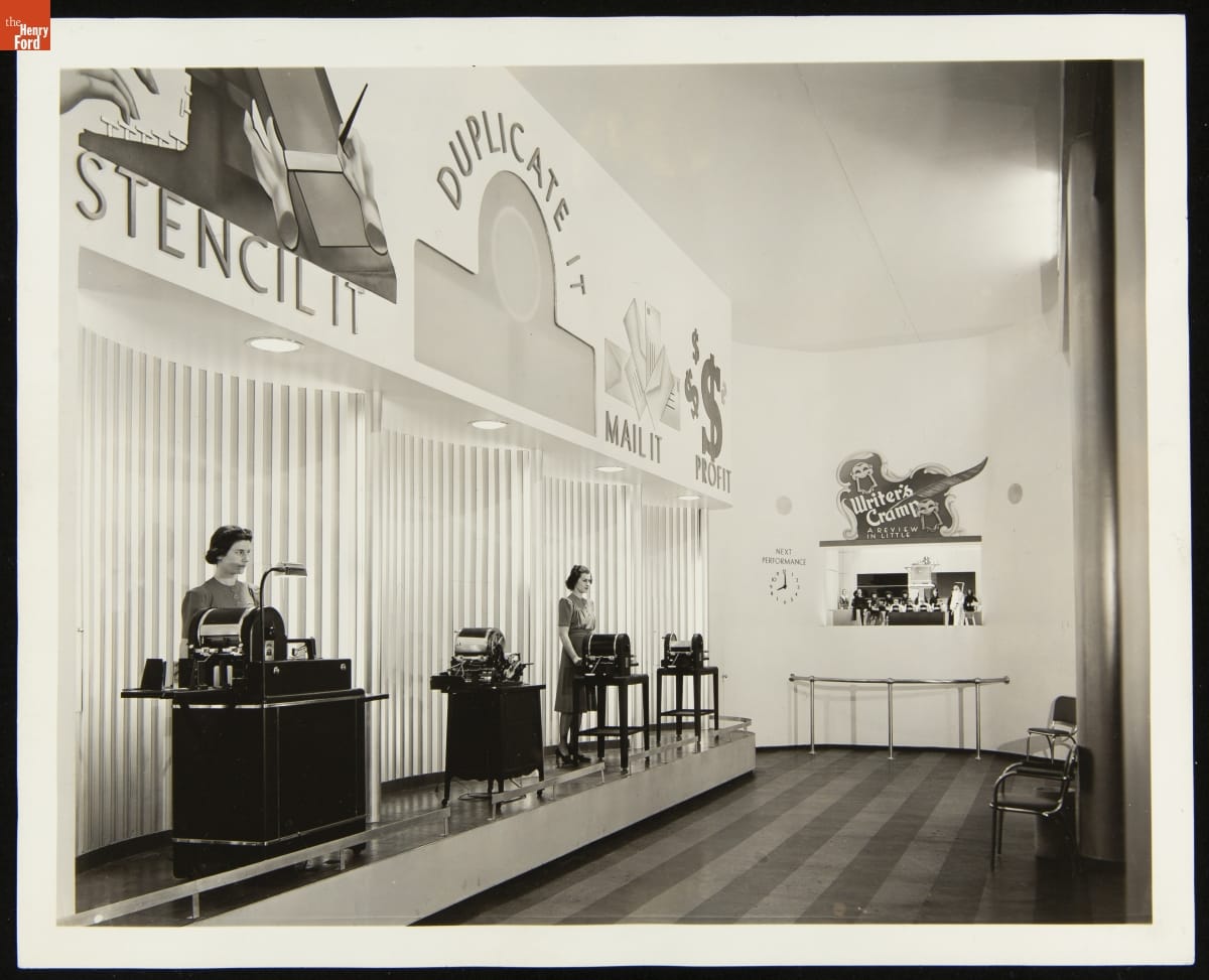 Room with display of office equipment and two women standing by it to the left, and puppet show in back