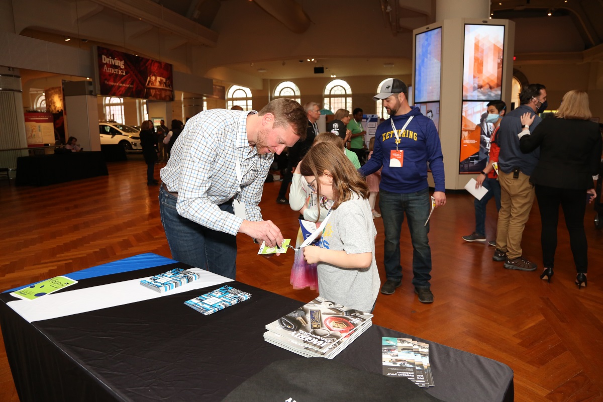 A man marks a child's lanyard in front of a table in a large space with other people and displays