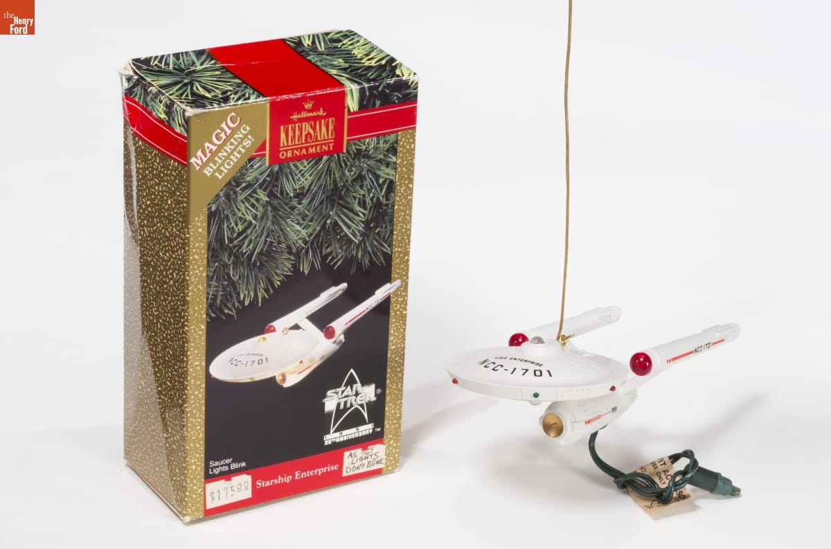 Ornament of spaceship with Christmas light extending from it, sitting next to gold-and-green box with image of the same ornament on the front