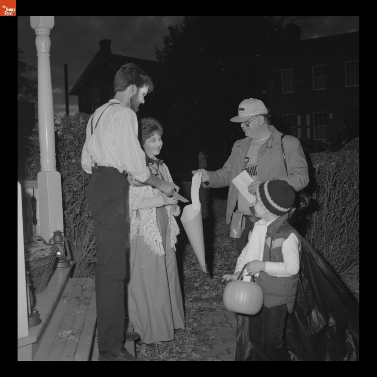 "Trick or Treat" at Wright Home in Greenfield Village, October 1982 Man and woman wearing historical clothing and facepaint hand candy to man and little boy