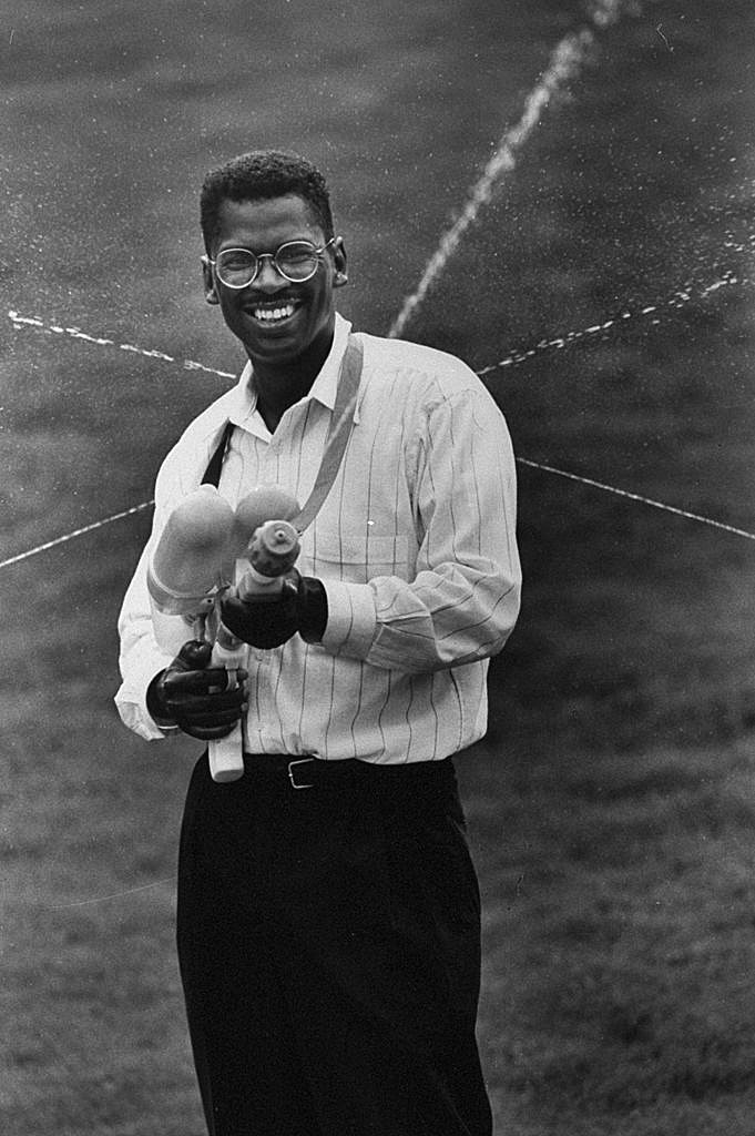 African American man wearing round glasses smiles and points large water gun toward camera