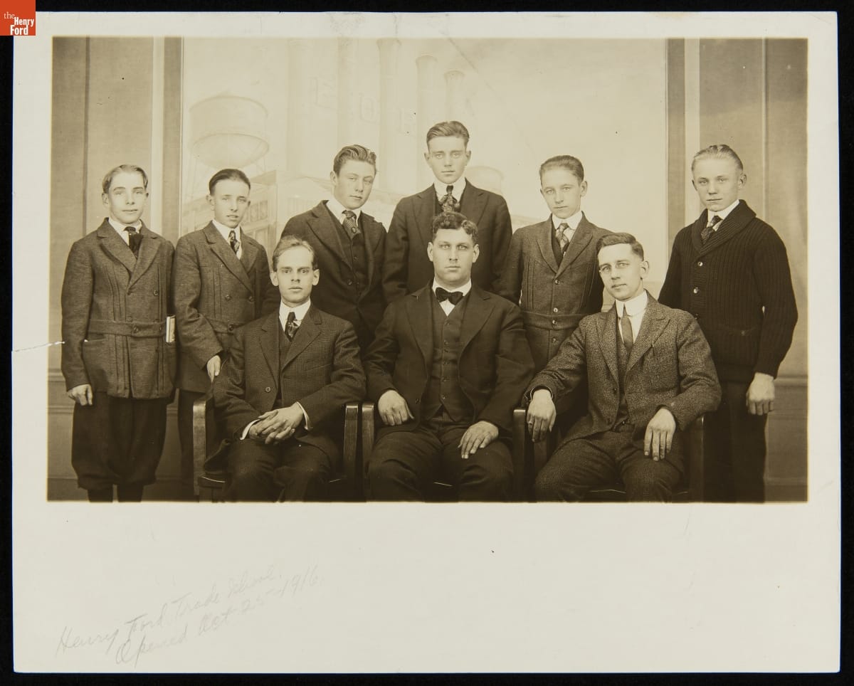 Group of nine sitting and standing boys and men, all wearing suits, pose for a photo