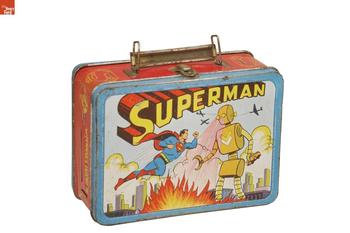 Lunchbox, Superman, 1954 Slightly rusted lunchbox with handle and front panel illustration of man in blue and red super hero outfit floating in air and fighting a large yellow robot shooting flames from its eyes; 3D text reading “Superman”