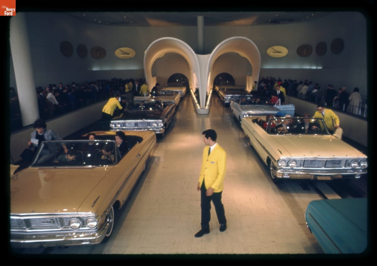 Assistants in yellow sports jackets assist people into convertible cars on two sets of tracks in a large room; more people queue on either side, waiting their turn