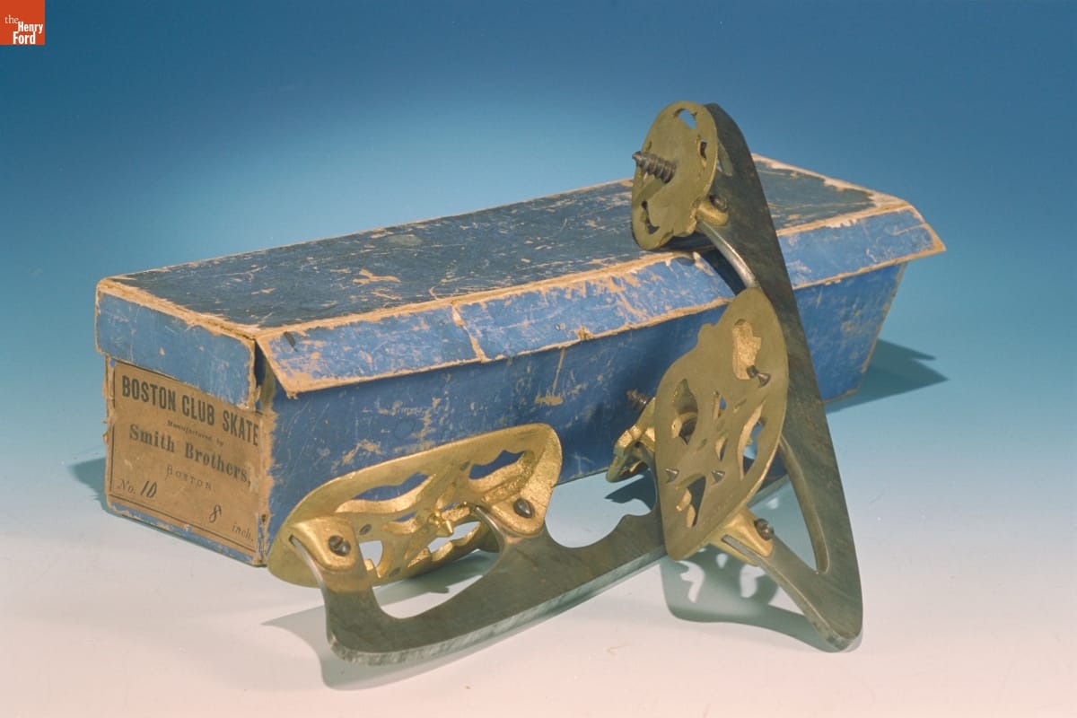 Blue shoebox with tan label on end and two gold and silver ice skating blades next to it