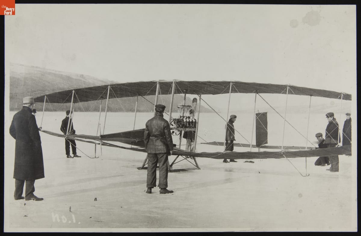 Black-and-white photo of an early glider on snow or ice with a number of people around it