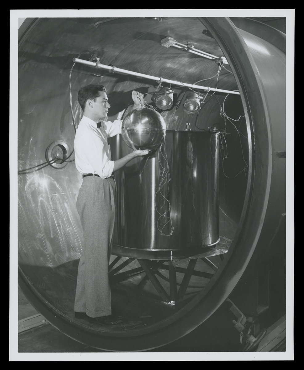 Man stands in large metal tube holding shiny sphere next to large vat