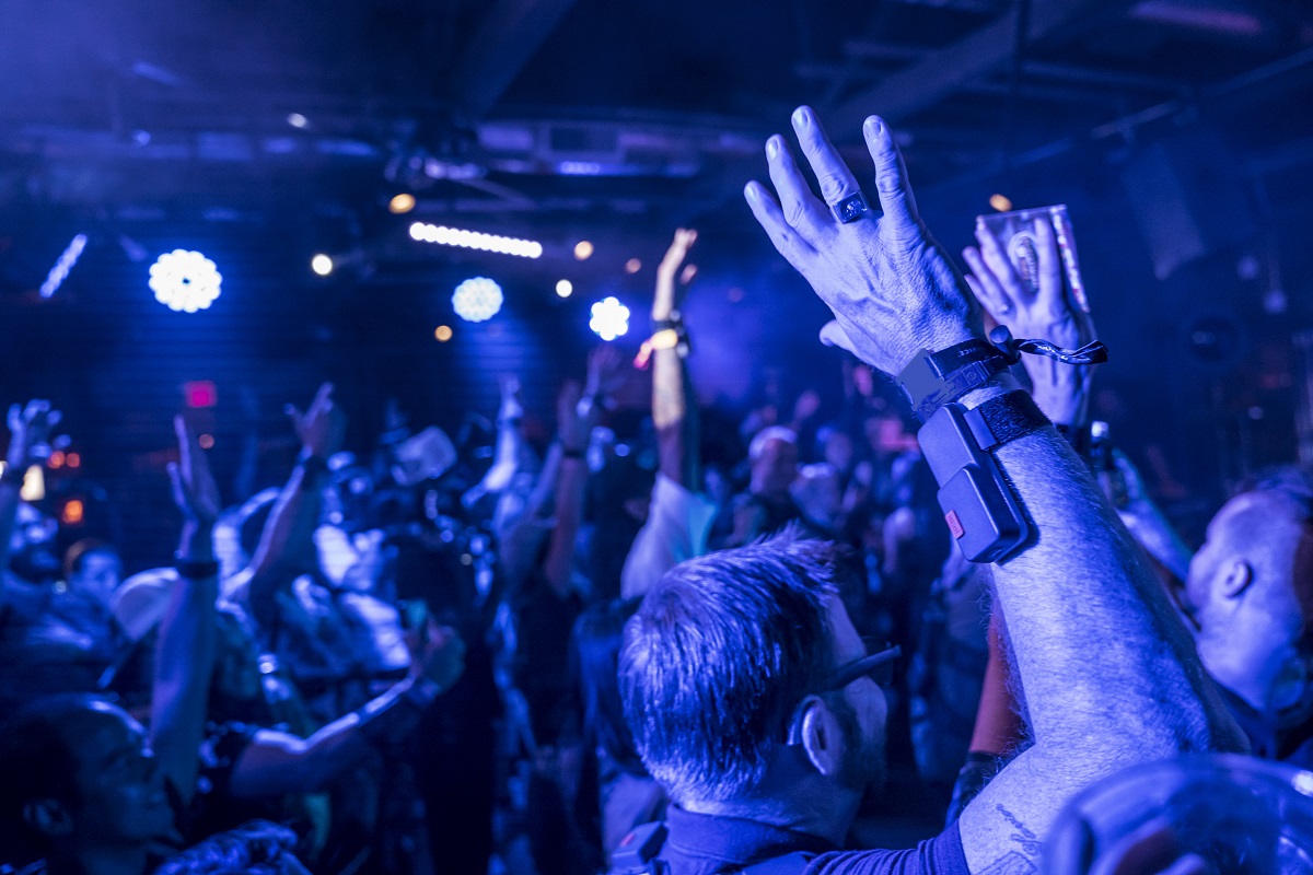 Room filled with blue light and people with their hands in the air; one close to camera wears a device strapped to his arm