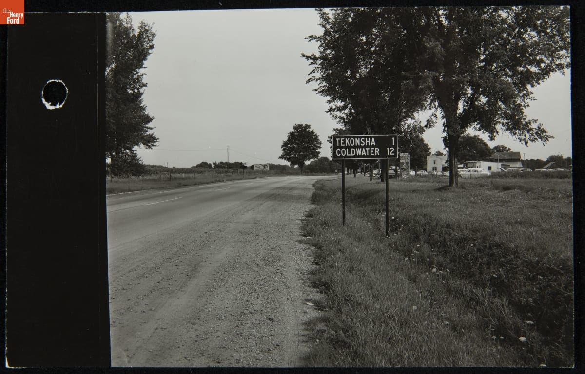 Road sign next to dirt road running by fields and buildings