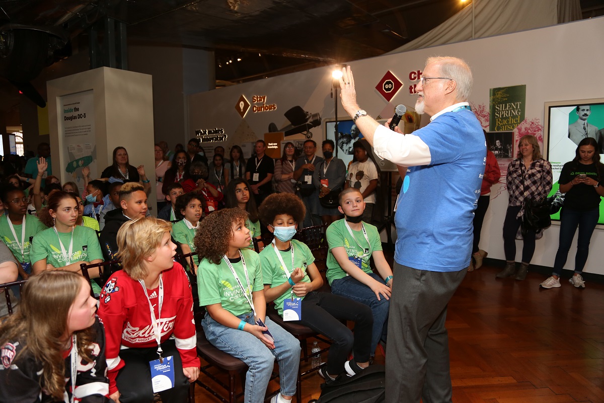 A man in a blue t-shirt talks to a group of children and adults