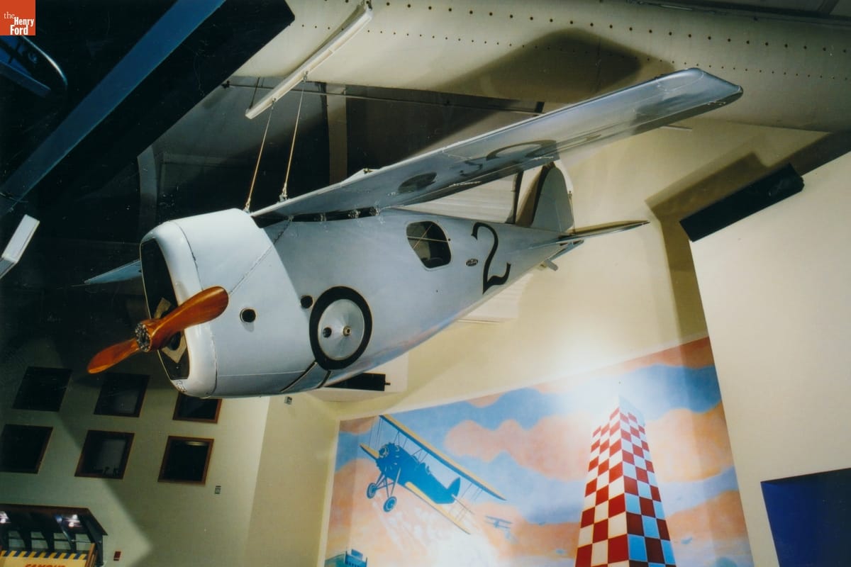 Small silver plane suspended from the ceiling in a room containing painted backdrops on the walls and other objects