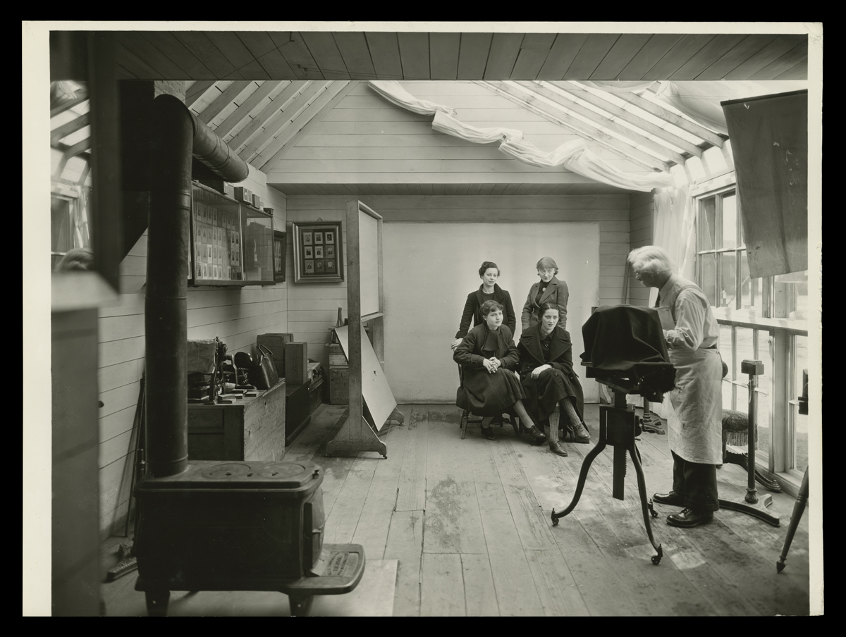 Room with photographer at camera pointed at four women posing; woodstove in foreground and images on walls