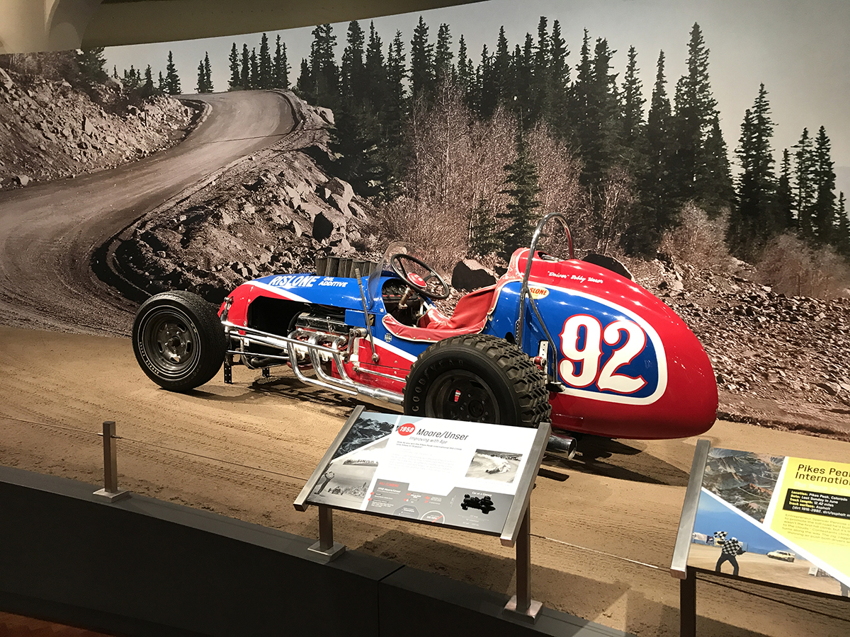 Red and blue race car with large number "92" on side sits on a dirt-like surface in front of a backdrop of a mountain road and trees; placards in front