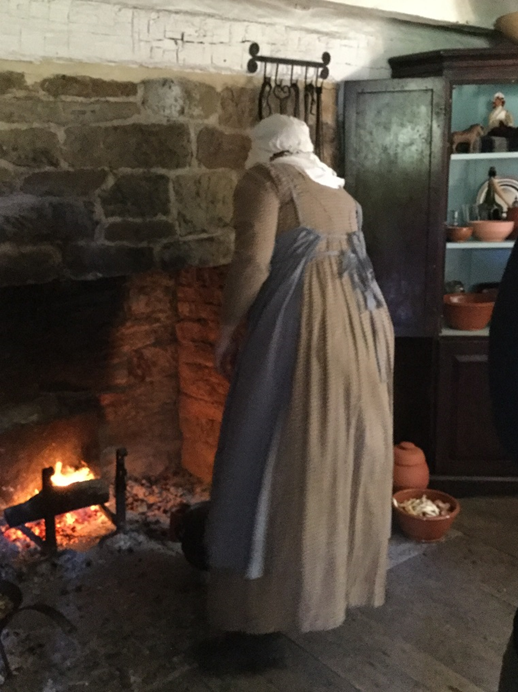 Woman in long simple dress and apron bends over a fire in a large stone fireplace